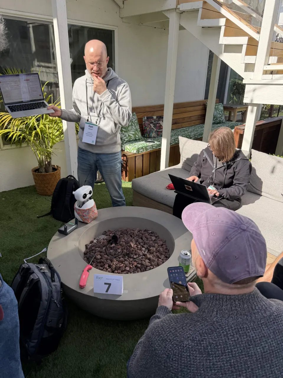 Ilan Strauss holds up a laptop at prototype table 7 while campers look on; a small white Reachy Mini robot sits on the edge of a fire pit
