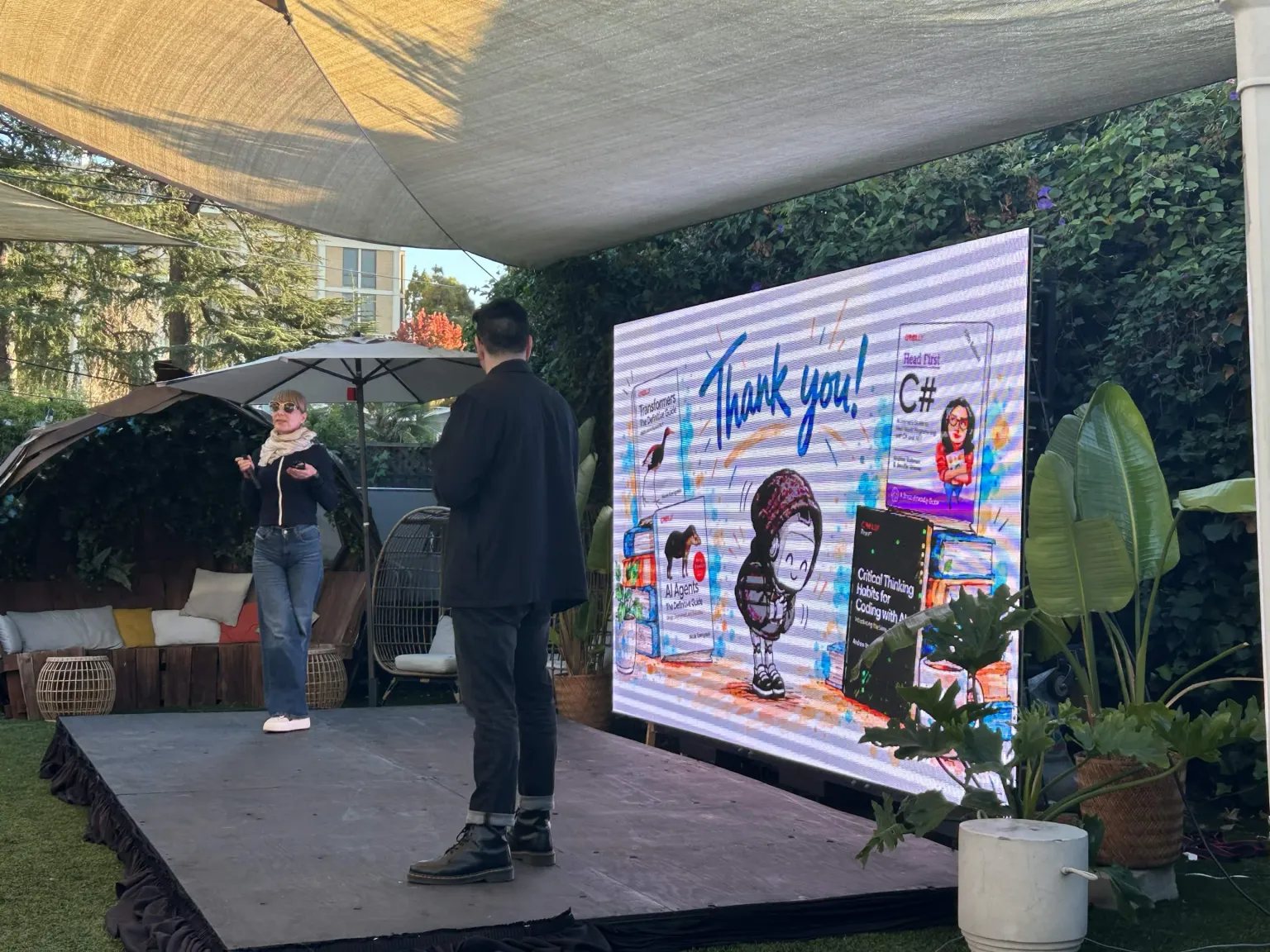A speaker on Lighthaven's outdoor stage, in front of a screen thanking campers and showing O'Reilly book covers